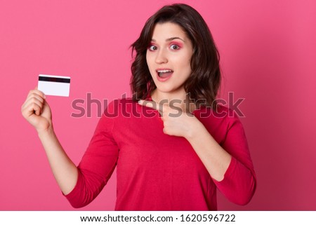 Picture of emotional good looking pretty female pointing with forefinger at credit card, having surprised facial expression, opening mouth, doing shopping online. Women and purchases concept.