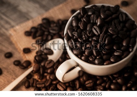 coffee photography background by selective focus at the roast coffee beans on a white coffee cup on a retro hemp sack and vintage dark wooden table decoration with blurred stainless steel coffee spoon
