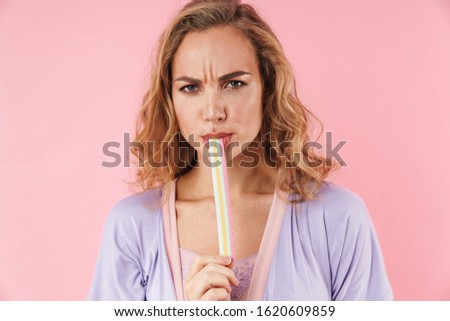 Image of caucasian displeased woman in stylish pajama eating marmalade isolated over pink background