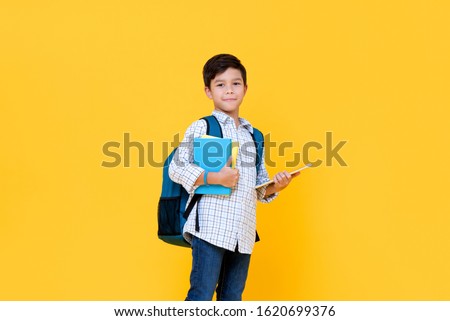 Handsome 10 year-old boy with backpack holding books and tablet computer in yellow background for education concept Royalty-Free Stock Photo #1620699376