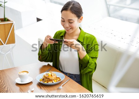 Smiling asian woman taking a photo of her pancakes for breakfast while sitting at the table indoors
