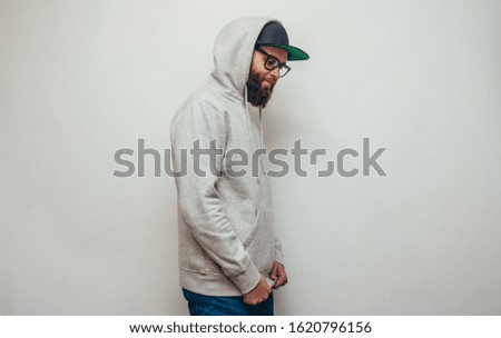 Handsome hipster guy with beard wearing gray blank hoodie or hoody and black cap with space for your logo or design on white background. Mockup for print
