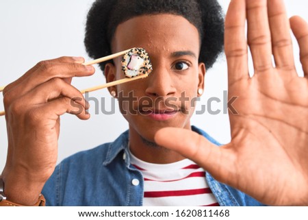 Young african american man eating sushi using chopsticks over isolated white background with open hand doing stop sign with serious and confident expression, defense gesture