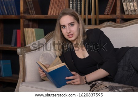 A girl (young woman) with blond hair reclines on her side, leaning on her elbow, and reads a book in a blue cover, in the background bookcases with books, the girl looks at the camera