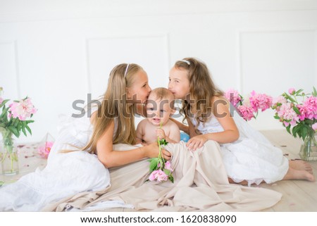 Cute little sisters with blond hair in white dresses and a little brother in a light spring studio decorated with pink and white peonies