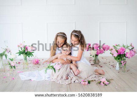 Cute little sisters with blond hair in white dresses and a little brother in a light spring studio decorated with pink and white peonies