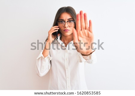 Young businesswoman wearing glasses talking on smartphone over isolated white background with open hand doing stop sign with serious and confident expression, defense gesture