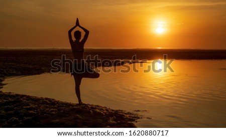 Vrikshasana asana. Young woman practicing tree pose at the beach during sunset golden hour. Arms raising with namaste mudra. Outdoor yoga. Sun reflection in water.  View from back. Melasti beach, Bali