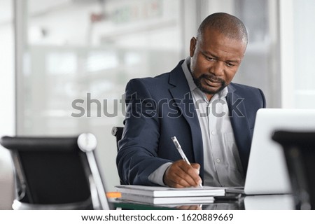 African serious businessman writing notes and using laptop. Mature business man writing his strategy in modern office. Focused black entrepreneur sitting at desk in modern office while working. Royalty-Free Stock Photo #1620889669