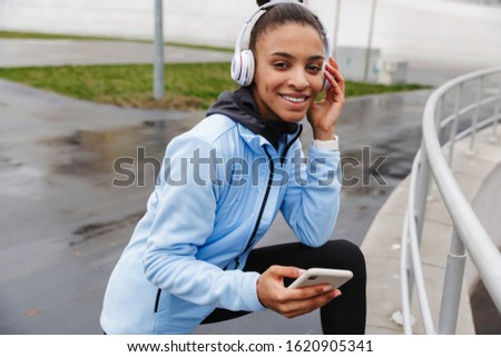 Attractive smiling young healthy african sportswoman listening to music with wireless earphones wrking out at the stadium, holding mobile phone