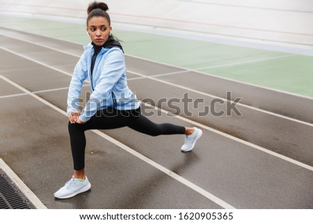 Beautiful young fit african sportswoman wearing wireless earphones doing stretching exercises at the stadium