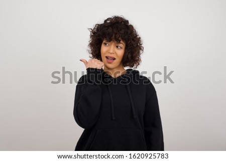 Omg concept. Stupefied caucasian female with surprised expression, opens eyes and mouth widely, points aside with thumb, shows something strange on gray background. Advertisement concept.