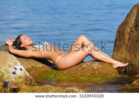 Young girl in white and blue swimsuit posing with eyes closed, sitting on large boulder near the sea