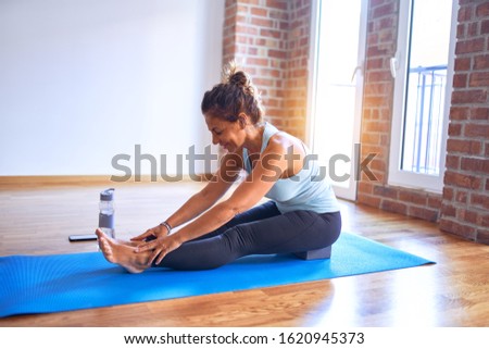 Middle age beautiful sportwoman smiling happy. Sitting on mat practicing yoga doing seated forward fold pose at gym
