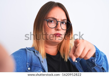 Beautiful woman wearing denim shirt and glasses make selfie over isolated white background pointing with finger to the camera and to you, hand sign, positive and confident gesture from the front