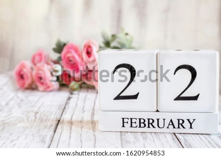 White wood calendar blocks with the date February 22nd. Selective focus with pink ranunculus in the background over a wooden table.