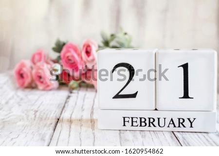White wood calendar blocks with the date February 21st. Selective focus with pink ranunculus in the background over a wooden table.