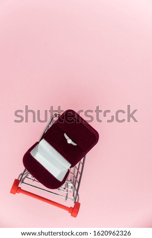Red box with wedding or engagement ring in a shopping cart on trendy pink background with copy space for text. Concept of holiday Valentine`s day shopping, sale and waste of money for love