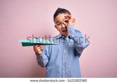 Young little boy kid holding paper plane over isolated pink background with happy face smiling doing ok sign with hand on eye looking through fingers