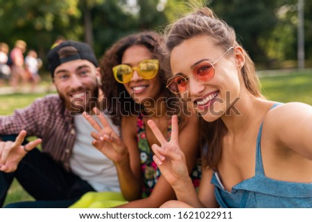colorful stylish happy young company of friends sitting park, man and women having fun together, summer hipster fashion style, traveling with camera, taking selfie pictures, smiling and positive