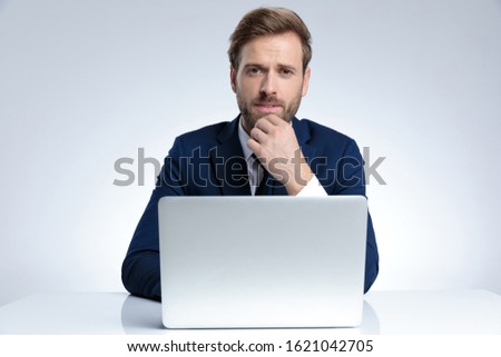 Bothered businessman biting his lips and disagreeing while holding his hand on his chin and sitting behind his laptop on gray studio background