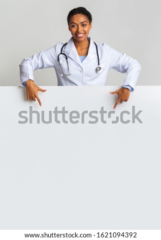 Free space for medical school ad. Smiling afro female intern doctor pointing at blank advertisement board with empty space, vertical shot