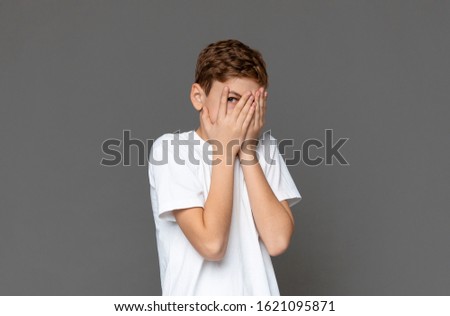 Shy teenage boy hiding, peeking through palms, grey studio background