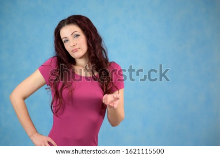 The model is smiling and talking cute to the camera. Portrait of a pretty slim brunette woman with emotions in a pink dress and with brown hair on a blue background in the studio.
