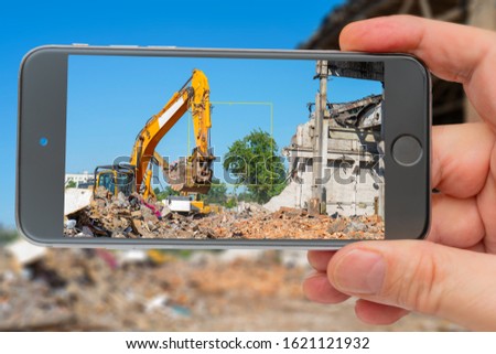Destruction of old building. Yellow excavator on ruins. Smartphone in hand. Digger on screen. 
