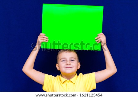Portrait of european schoolboy in yellow shirt posing with blank paper against blue background.