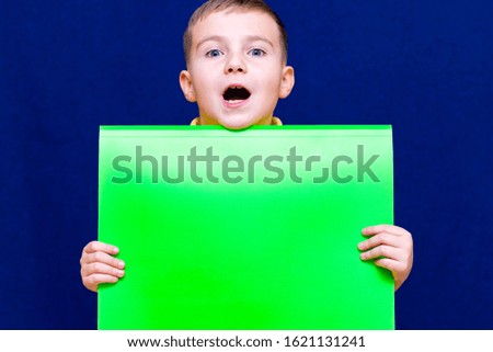 Attractive caucasian schoolboy posing with blank paper against blue background.Boy is opened mouth.