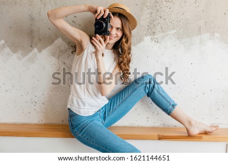 young pretty smiling happy woman taking pictures on vintage photo camera wearing shirt sitting against wall in straw, traveler in summer outfit, fashion trend