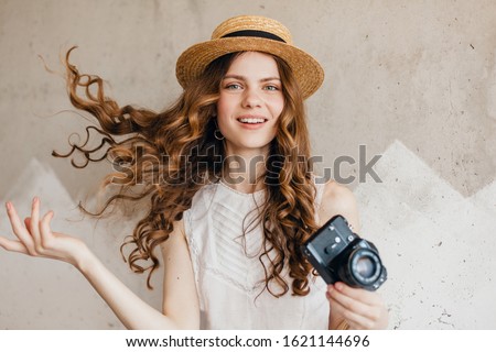 young pretty smiling happy woman wearing white blouse sitting against wall in straw hat holding vintage photo camera, traveler in summer outfit, fashion trend, waving long curly hair