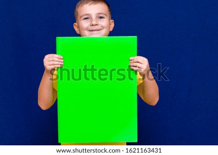 Beautiful european schoolboy in yellow shirt posing with blank green paper against blue background.