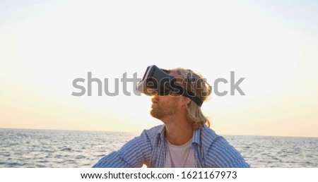 Portrait of young handsome male on the beach wearing virtual reality glasses, beautiful sunrise on the background 
