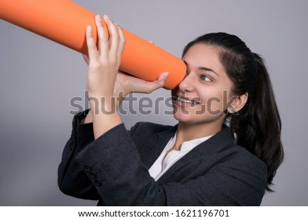 Indian young business woman in a business suit, holding a twisted orange tube in her hand in her hand, looking into it. Free space. Studio photography. On a gray background.