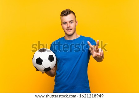 Young handsome blonde man holding a soccer ball over isolated yellow background smiling and showing victory sign