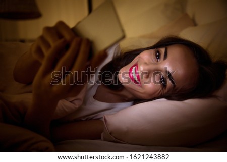 A night owl. Close-up photo of an insomniac girl, who is surfing the Internet on her smartphone at night, lying alone in her bed and smiling.