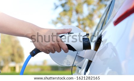 Electric car or ev is charging at station . man use the white power cable and plug on nature background. eco and clean energy concept with the mobile phone smart technology. zero gas no transmission Royalty-Free Stock Photo #1621282165