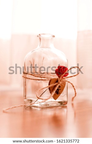 Close up image of transparent glass bottle with a heart tied with string and red rose, blurred background in sepia tones.