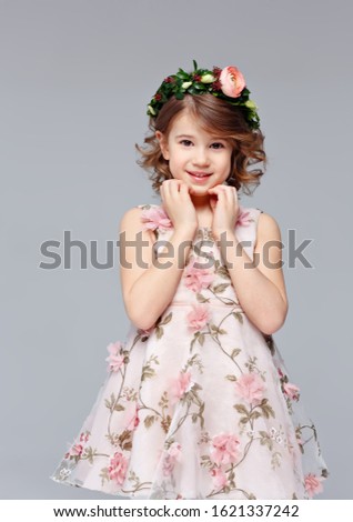 Portrait of a beautiful cut little girl in an elegant dress with a wreath of fresh flowers on her head stands, lauht and looks into the camera in the studio on a gray background.
