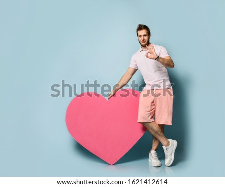 Young handsome man hold pink heart shape toy present gift in hand and show cool good ok sign on pastel blue wall background. Love gift for valentines day with text space