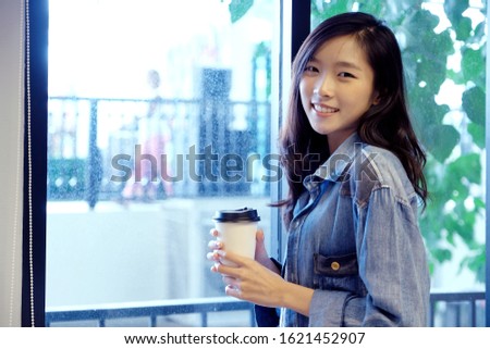 Asian woman with take away coffee cup, Happy asia girl looking at camera and holding coffee while standing by window,  Asian female university student and disposeble cup in coffee shop cafe background