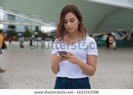 Front view of calm woman holding smartphone. Beautiful young lady using modern phone while standing on street. Technology concept