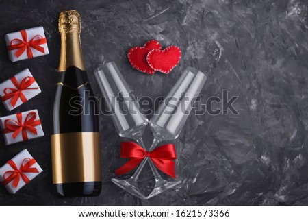 Top view of a champagne bottle with a blank label, two glasses with a red satin ribbon bow, two handmade hearts and four gift boxes with copy space on a textured black background.