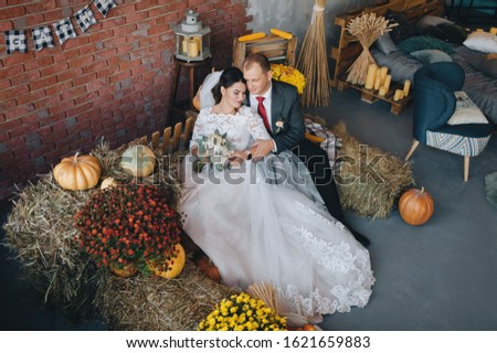 Stylish groom and beautiful bride in a white dress with a bouquet are sitting on the hay in the studio with autumn decor. Wedding portrait of happy and smiling newlyweds. Photography, concept.