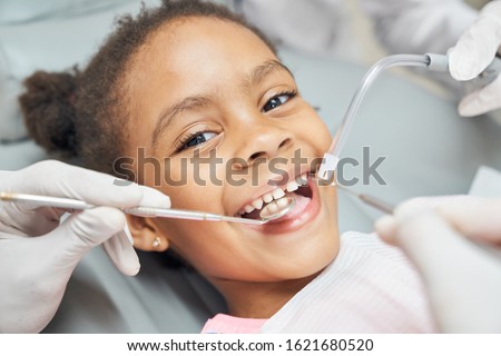 Charming little afro american girl sitting in dental chair, smiling and looking at camera during medical treatment at modern clinic. Concept of health care ad pediatrics Royalty-Free Stock Photo #1621680520