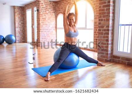 Middle age beautiful sportswoman smiling happy. Doing exercise using fitness ball in a class of yoga at gym