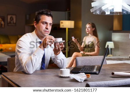 Elegant man sitting at table with laptop and cup of coffee while his wife relaxing in home interior