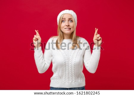 Young woman in sweater, hat isolated on red background in studio. Healthy fashion lifestyle cold season concept. Mock up copy space. Waiting for special moment, keeping fingers crossed, making wish